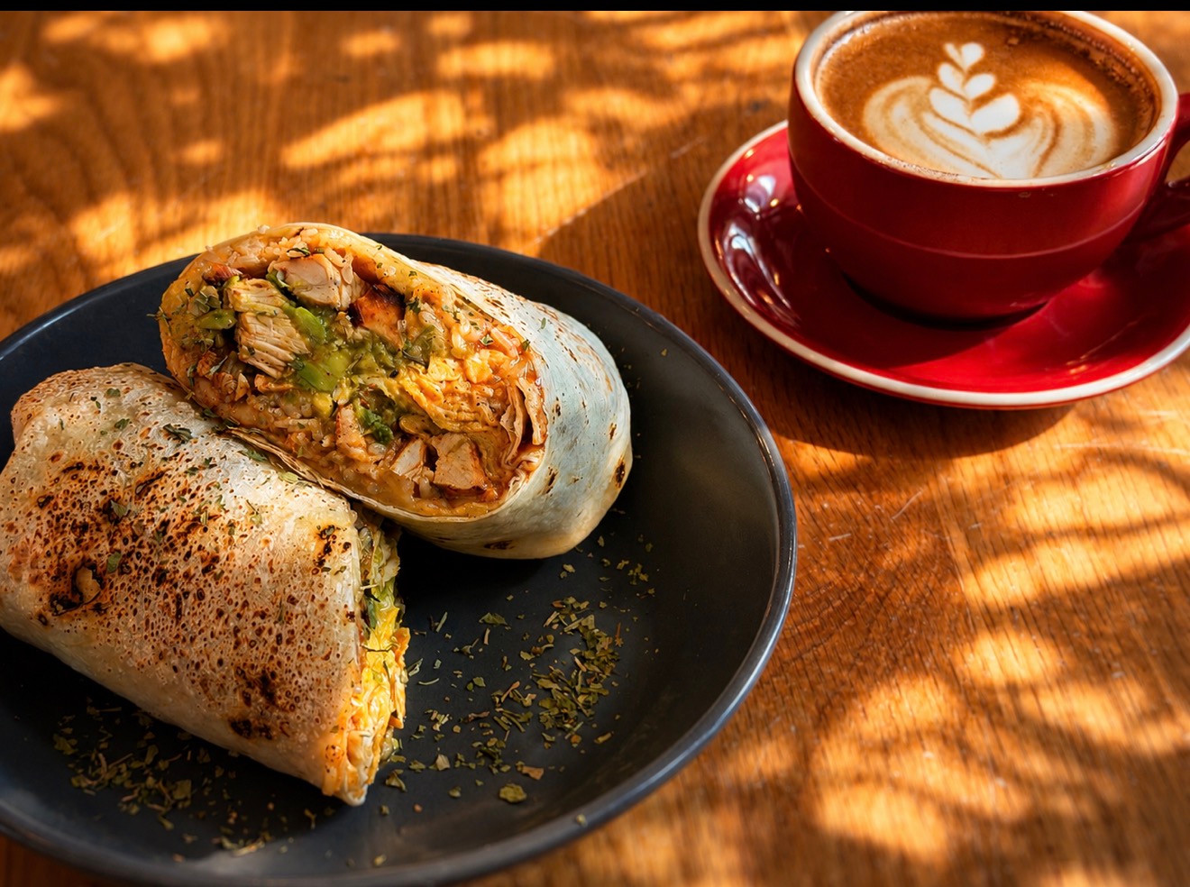 A toasted burrito cut in half on a dark plate beside a red cup of coffee.