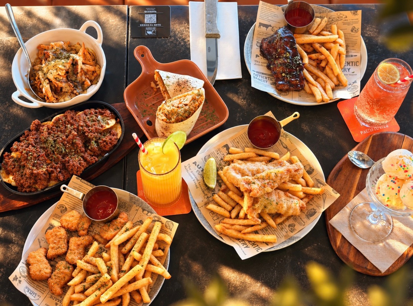 An overhead table spread with fish and chips, ribs, a burrito, pasta, colourful drinks and ice cream.