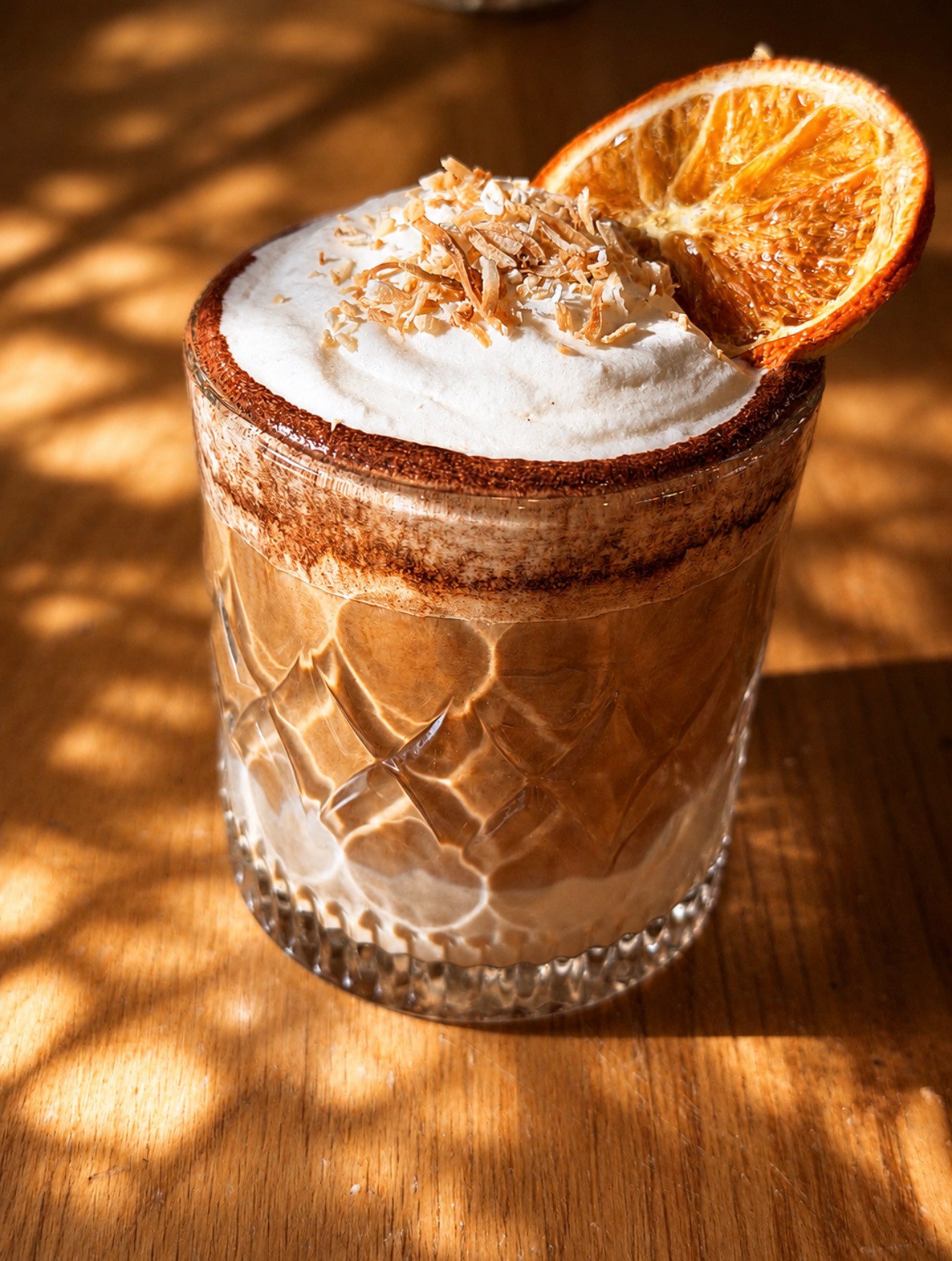 A cream-topped brown drink garnished with a dried orange slice on a sunlit wooden table.