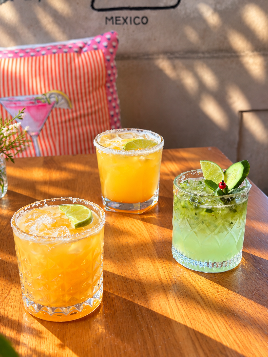 Two orange drinks and a green drink garnished with lime and cucumber on a sunlit wooden table.