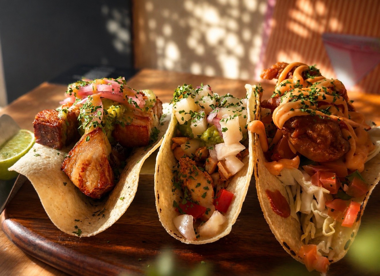 Three filled tacos topped with herbs, onions and sauce on a wooden serving board, with a lime wedge.