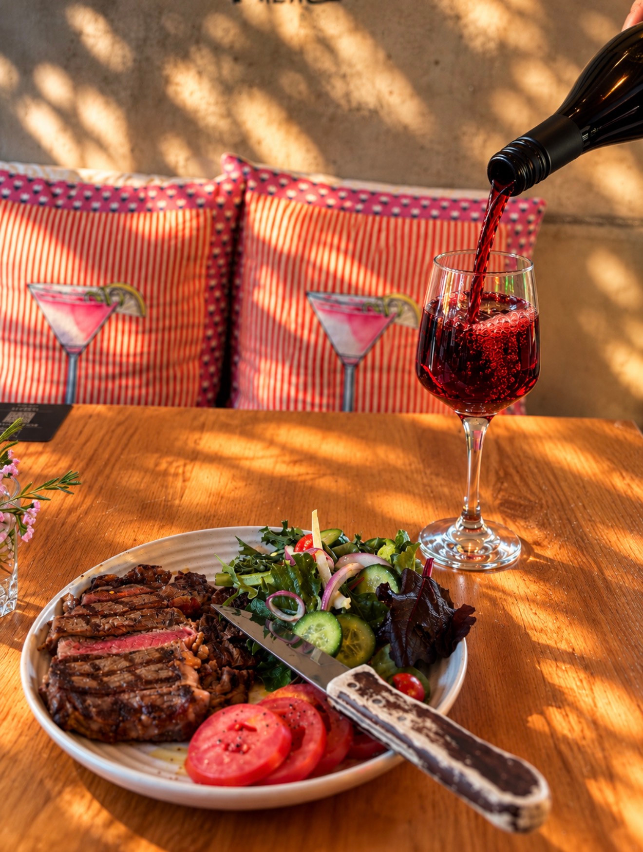 Red wine being poured into a glass beside sliced steak, salad and tomatoes on a sunlit table.
